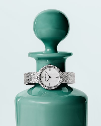 Image of Joia de Baume & Mercier watch on green vase. Created for the modern woman who blends refinement with fierce independence. Tap to shop the brand image